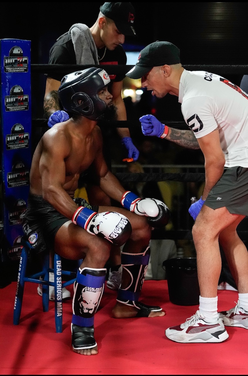 Fighter in gloves and shin guards seated in the corner while a coach talks him through the next round